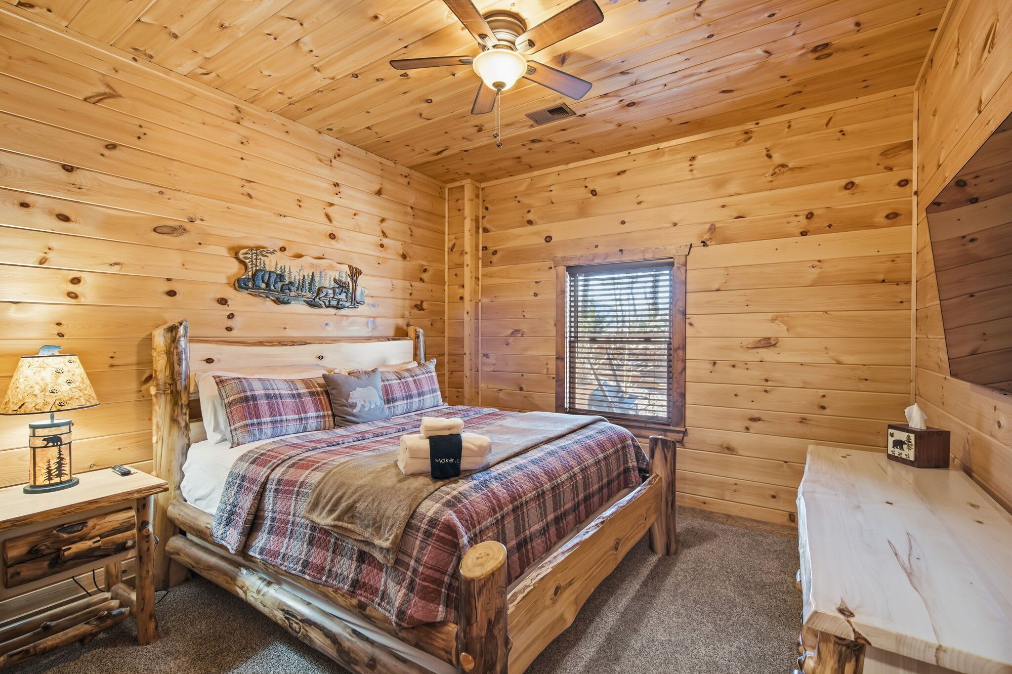 A wooden cabin room with a bed, a nightstand, a lamp, a window with blinds, and a ceiling fan.