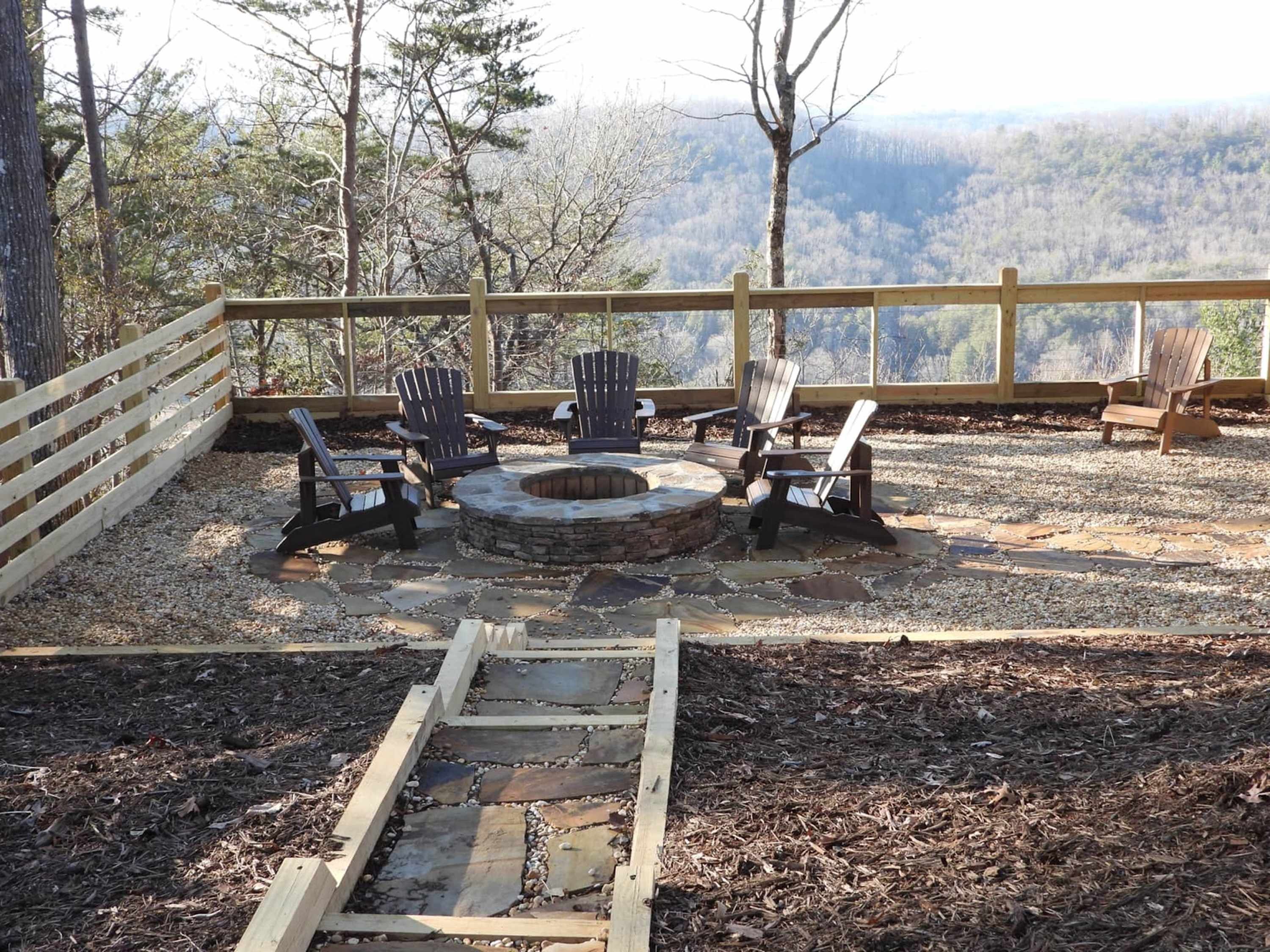 Een terras buiten met Adirondack-stoelen, een vuurkorf van natuursteen en een houten vlonder met hekwerk, uitkijkend over een bebost landschap.