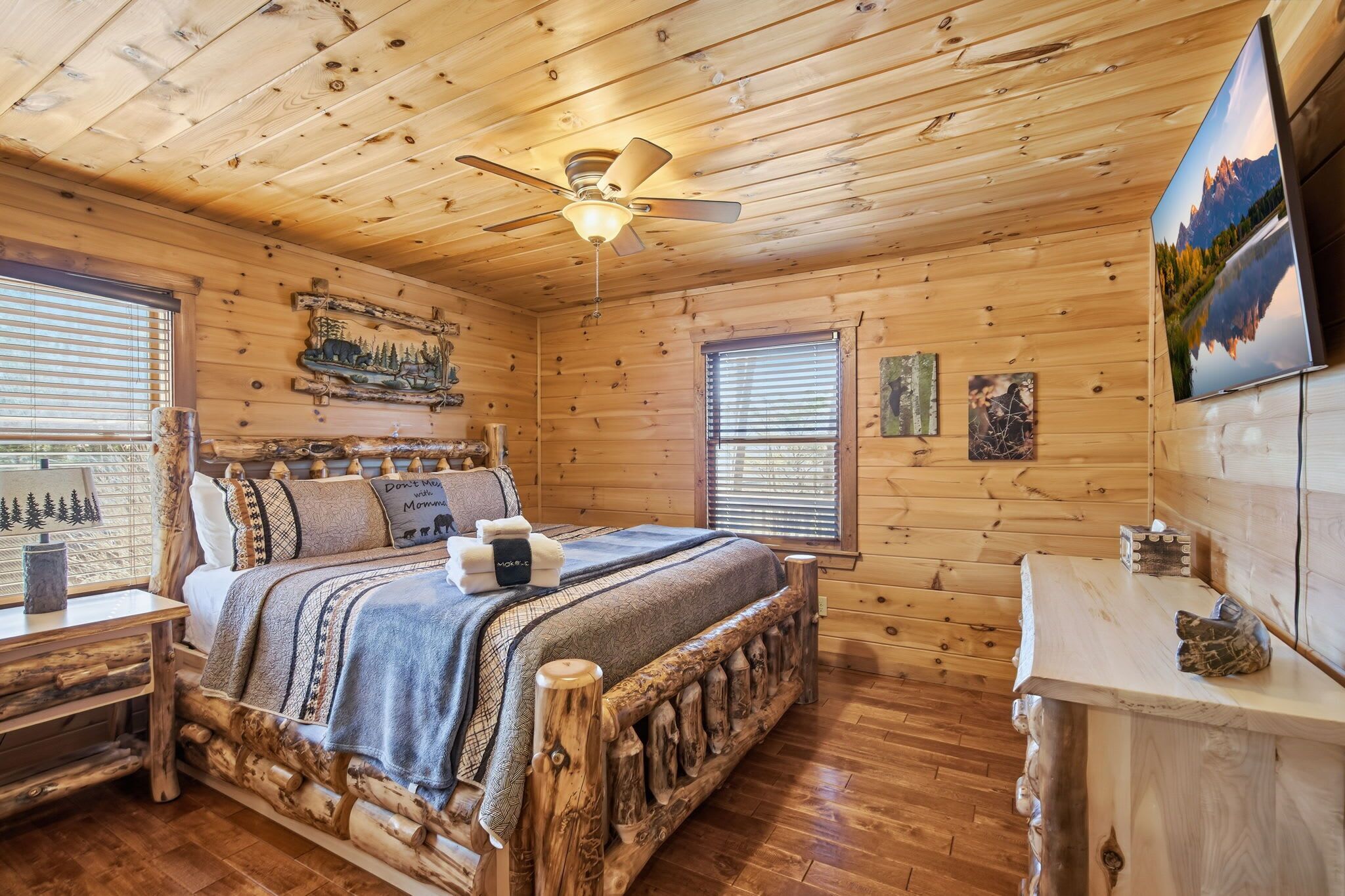 Un dormitorio tipo cabaña de madera con una cama de madera, un ventilador de techo y un ventanal grande.