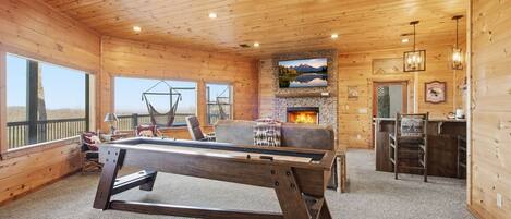 Interior de una cabaña de madera con mesa de billar, chimenea y vistas al exterior.