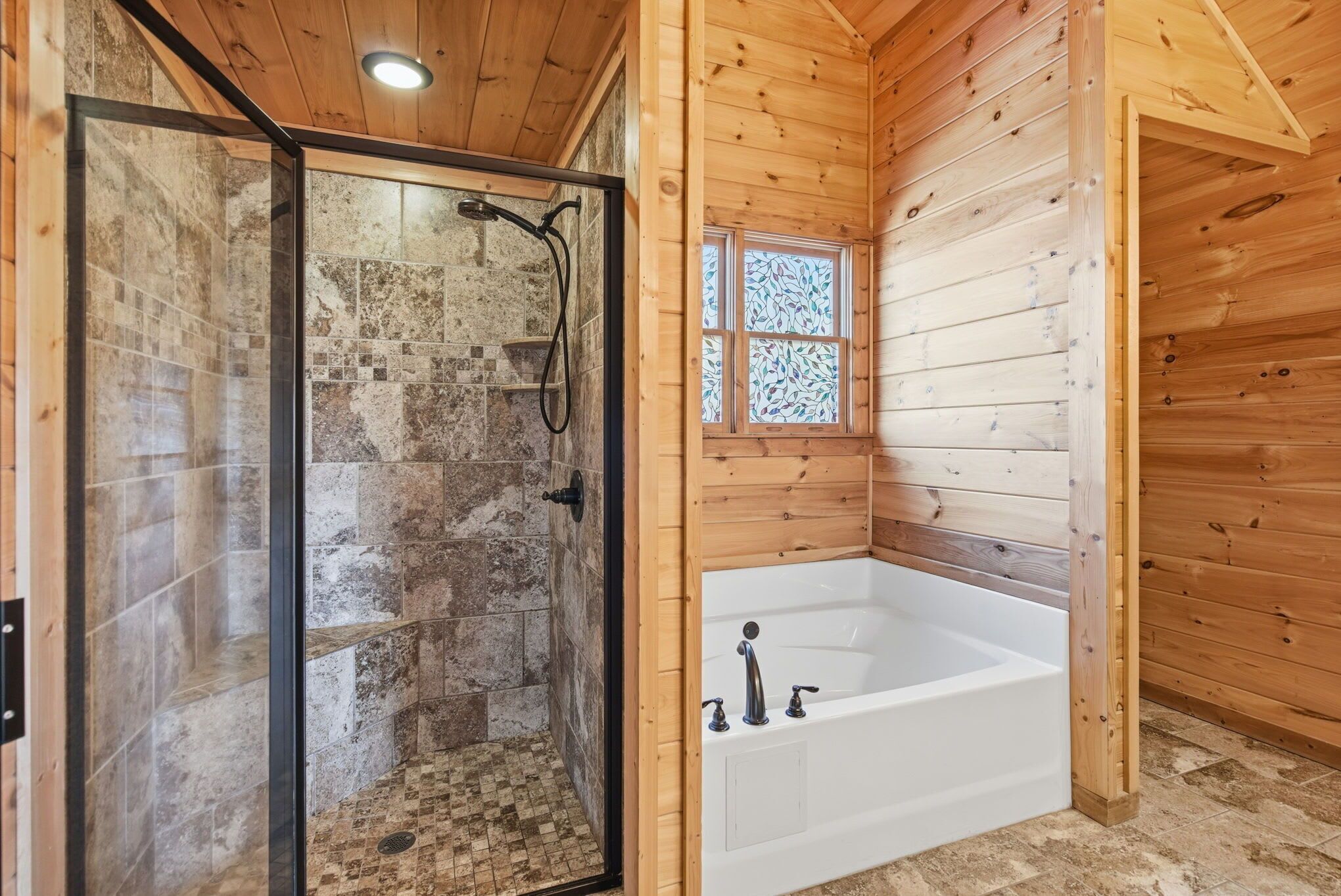 Baño de cabaña de madera con ducha envuelta en vidrio, una bañera blanca y una pequeña ventana con vitrales.