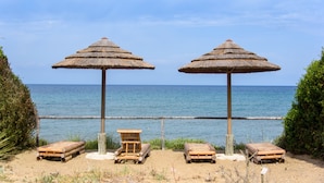Dos sombrillas de paja, dos sillones de madera y una pequeña mesa de madera sobre una playa arenosa, con el océano al fondo.