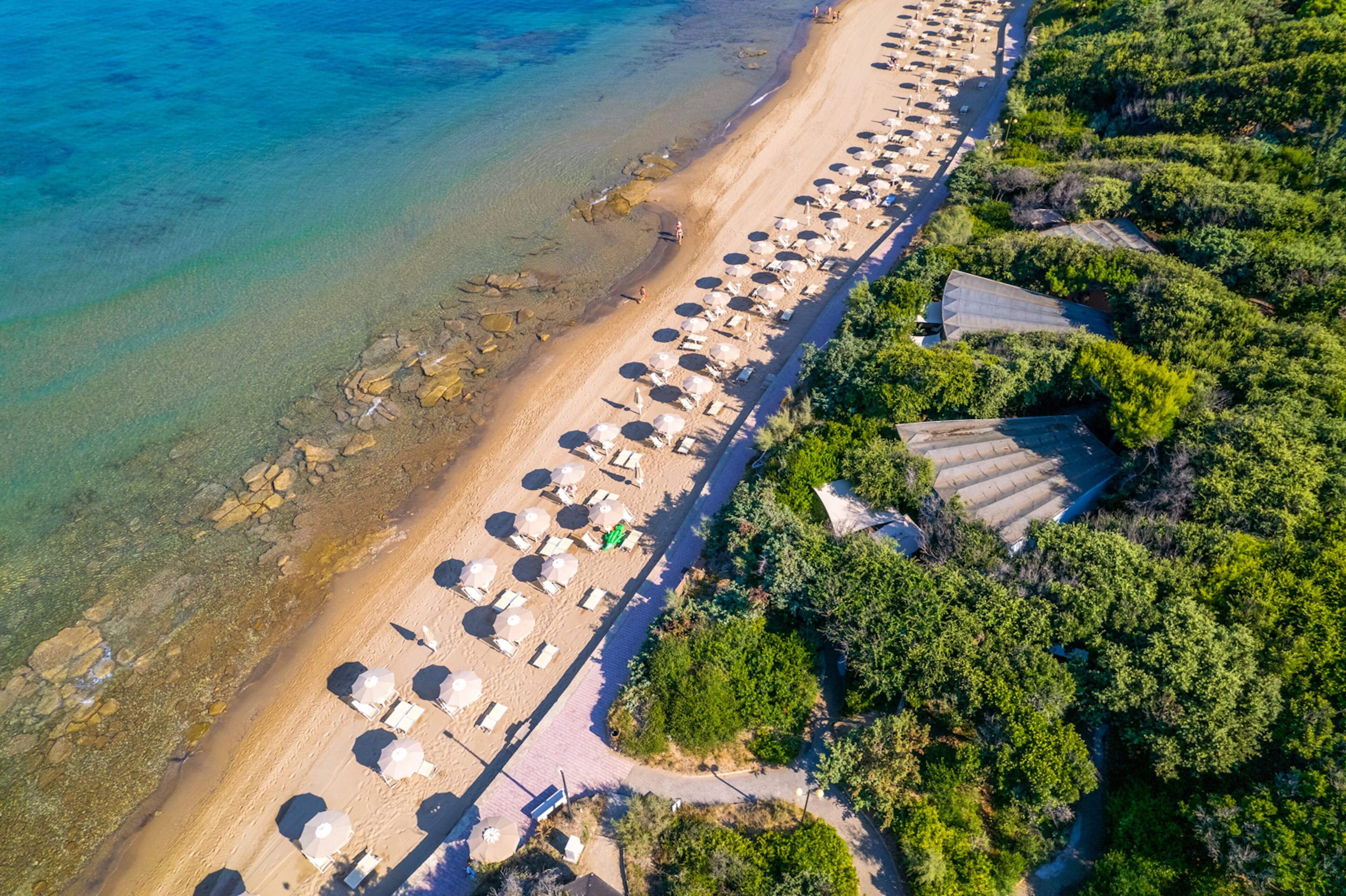 An aerial view of a beach with rows of sun loungers and umbrellas, a clear blue sea, and lush greenery.