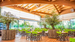 Amplio espacio interior con vigas de madera y ventanas grandes, con sillas y mesas verdes dispuestas para comer.