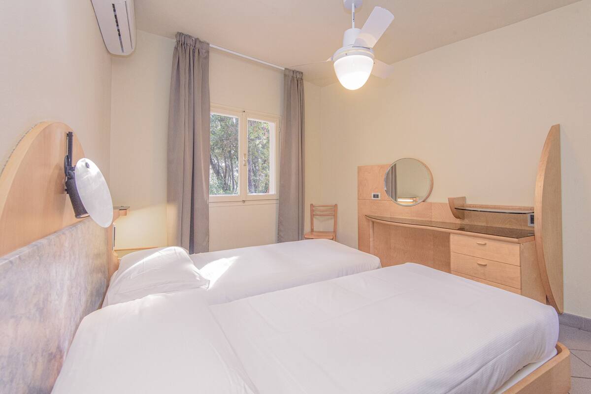 A hotel room with two beds, a ceiling fan, a window with curtains, a wooden desk with a mirror, and a small chair.