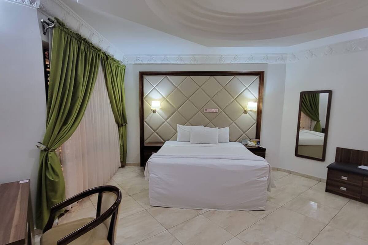 Ein Hotelzimmer mit Bett, Nachttisch, Sessel, Spiegel und grünen Vorhängen.