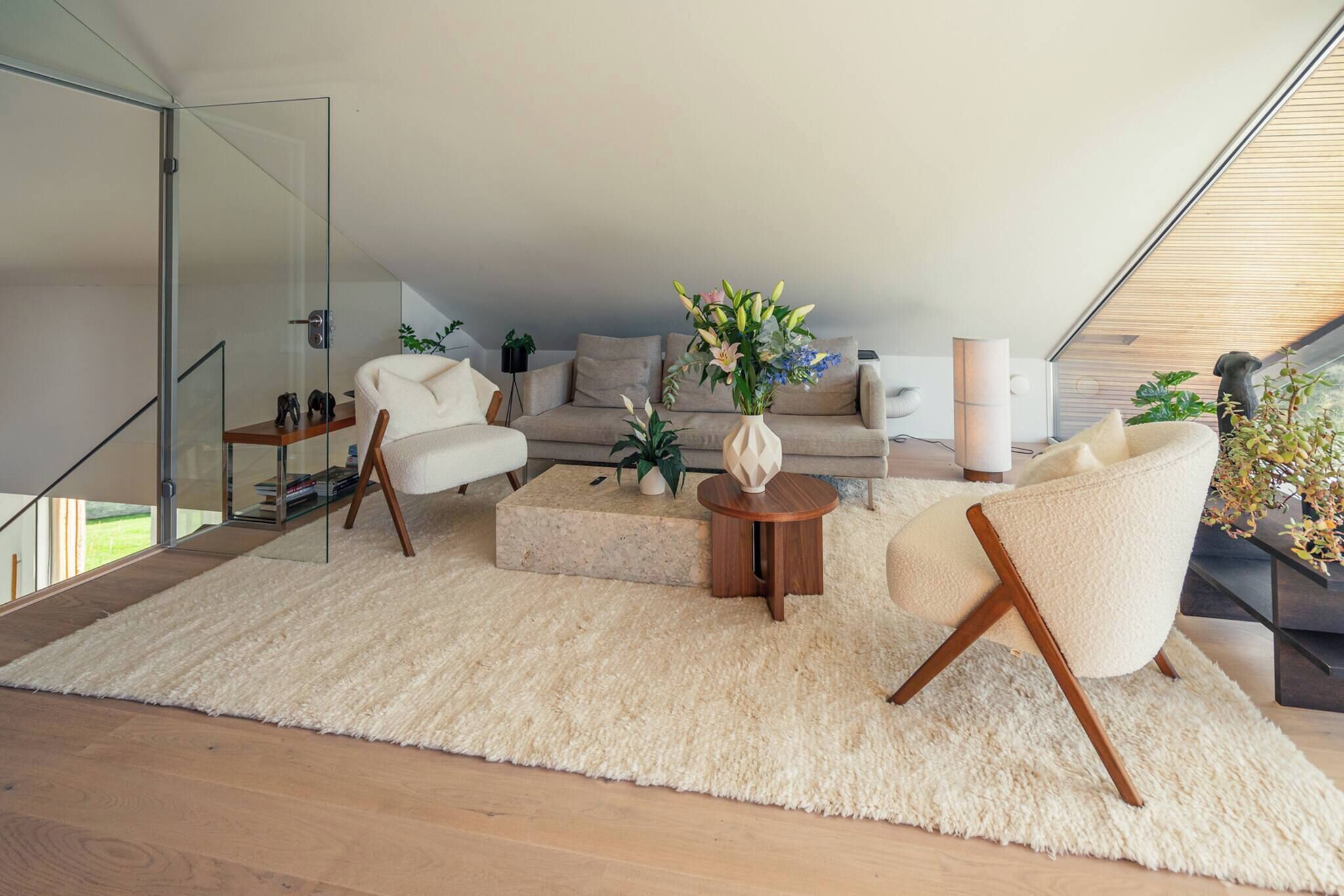 A modern living room with a sofa, two armchairs, a coffee table, and a rug. There is a glass door leading to a balcony, and a small table with a plant.