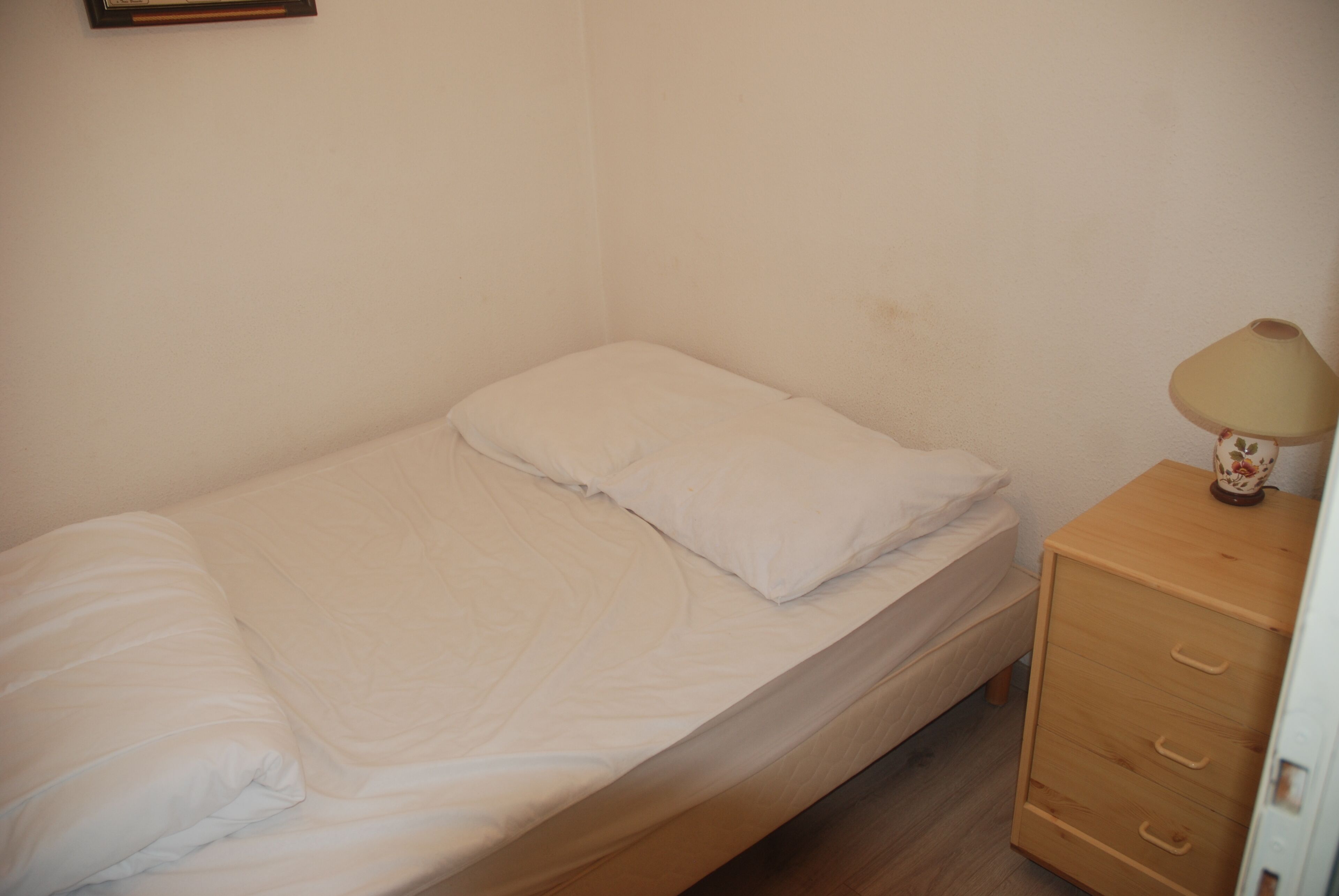 Ein kleines Zimmer mit einem Einzelbett, einem Holznachttisch und einer Lampe.