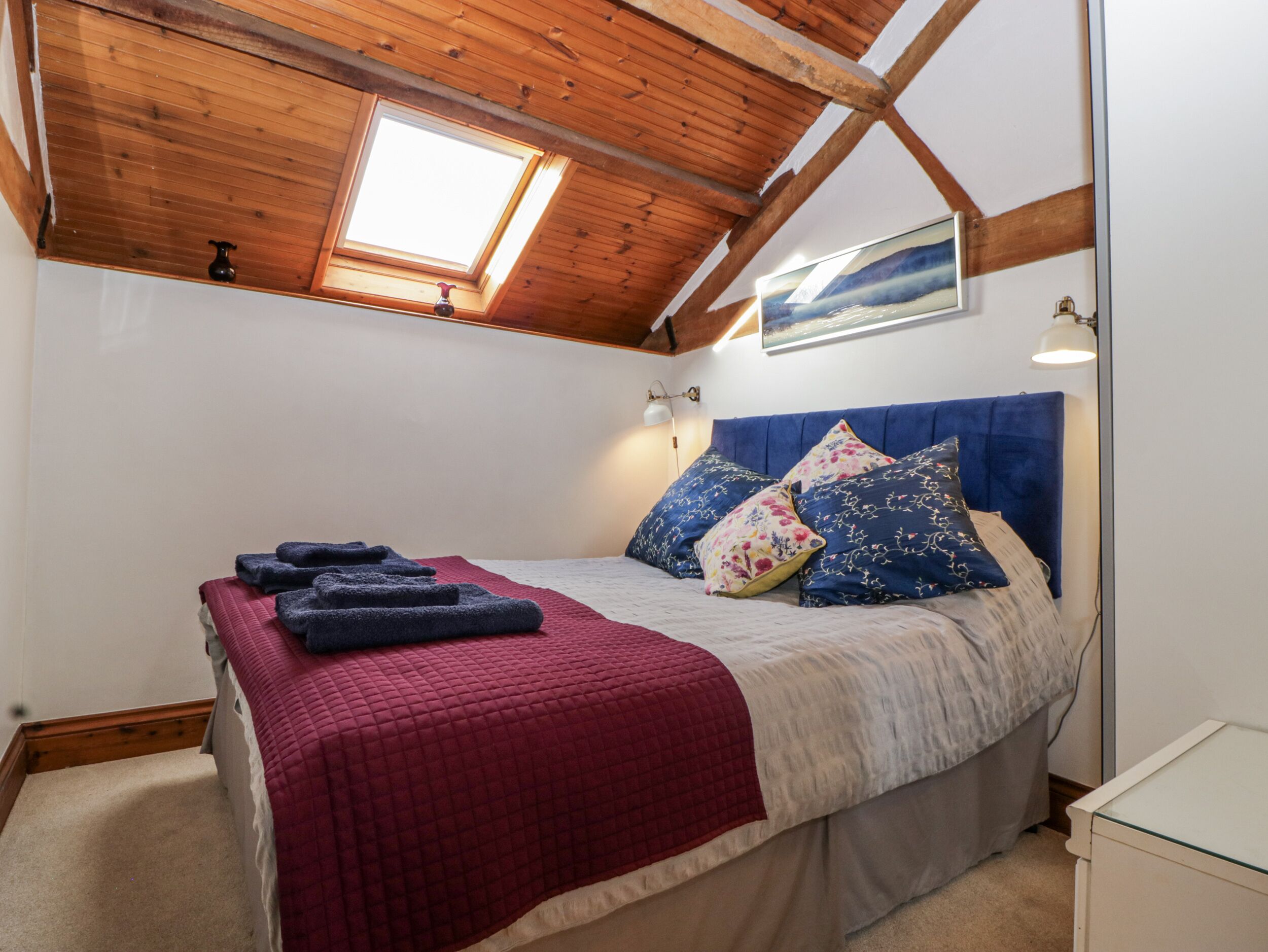 Une chambre avec un plafond en pente, un lit recouvert d’une couverture rouge, des coussins bleus à motifs floraux et un tableau accroché au-dessus du lit.