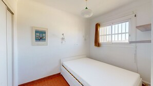 A small, clean room with a single bed, a window with a curtain, a wall-mounted light fixture, and a framed picture. - Seafront apartment for 4 people (Canet-en-Roussillon)