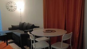 Una zona giorno accogliente con un tavolo rotondo e quattro sedie, un divano, una sveglia appesa al muro e una tenda.