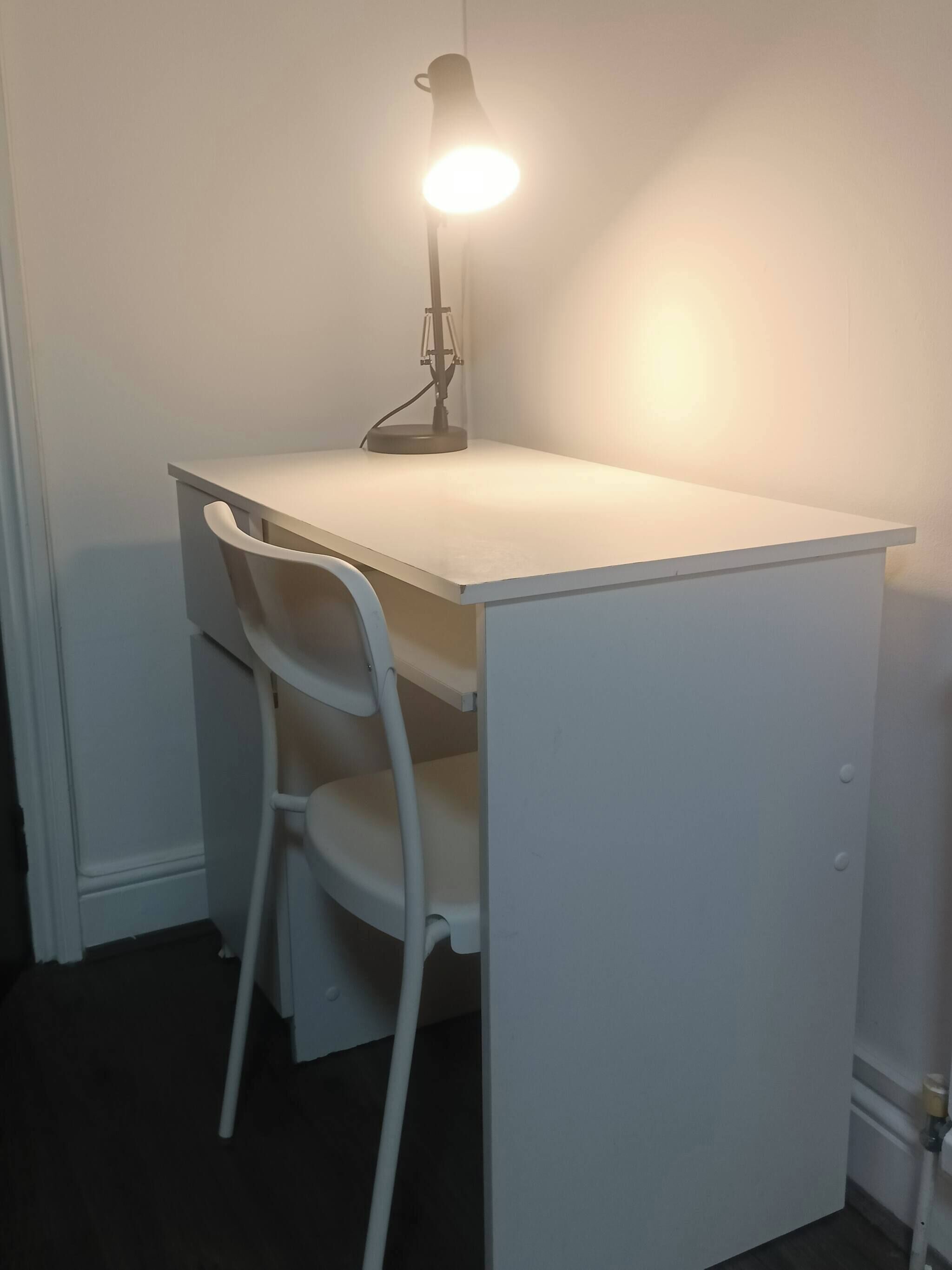 A white desk with a lamp, a white chair, and a closed cabinet.