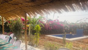 Un área de comedor al aire libre con techo de paja, rodeada de abundante follaje y plantas en flor.