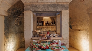 Habitació amb parets de pedra, una taula on hi ha diversos aliments i una estanteria amb ampolles de vi.