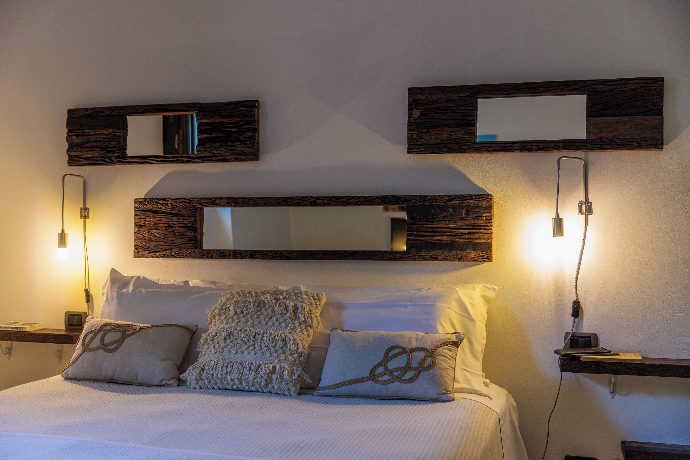 Una cama bien tendida con sábanas blancas y almohadas decorativas, un espejo montado en la pared y lámparas de noche.