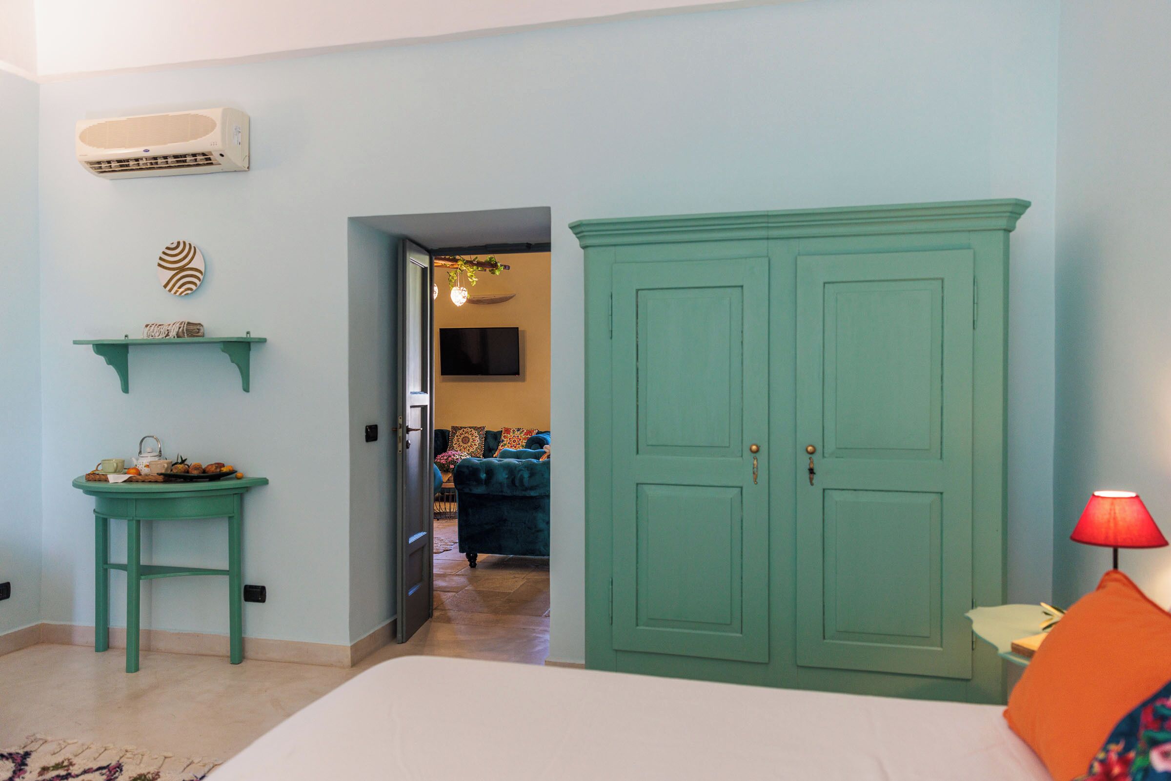 Una habitación con una cama, un armario verde, una mesita con lámpara y un aire acondicionado instalado en la pared.