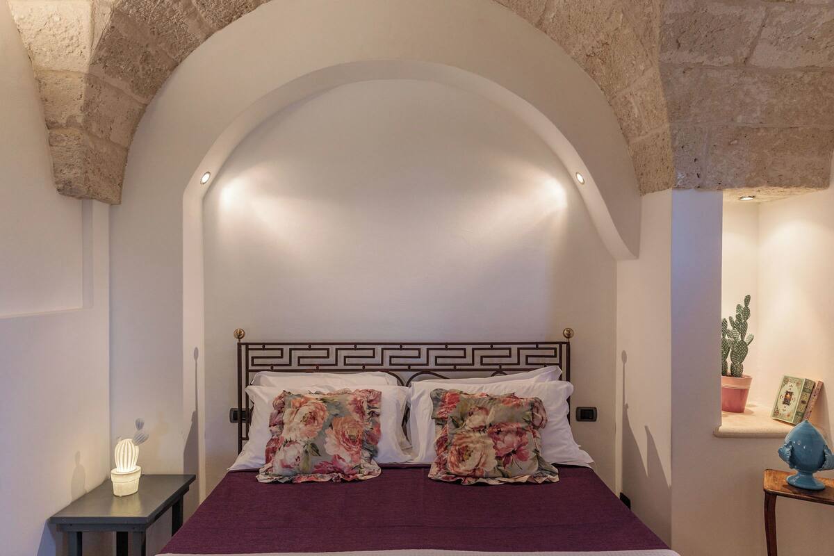 A bedroom with a stone arch ceiling, a bed with floral pillows, a bedside table with a lamp, and a cactus on a shelf.