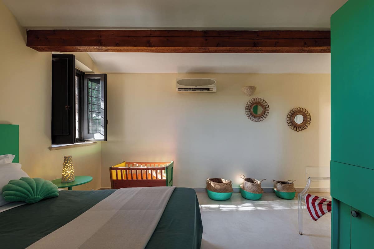 A bedroom with a bed, a crib, a window, an air conditioner, and decorative baskets.