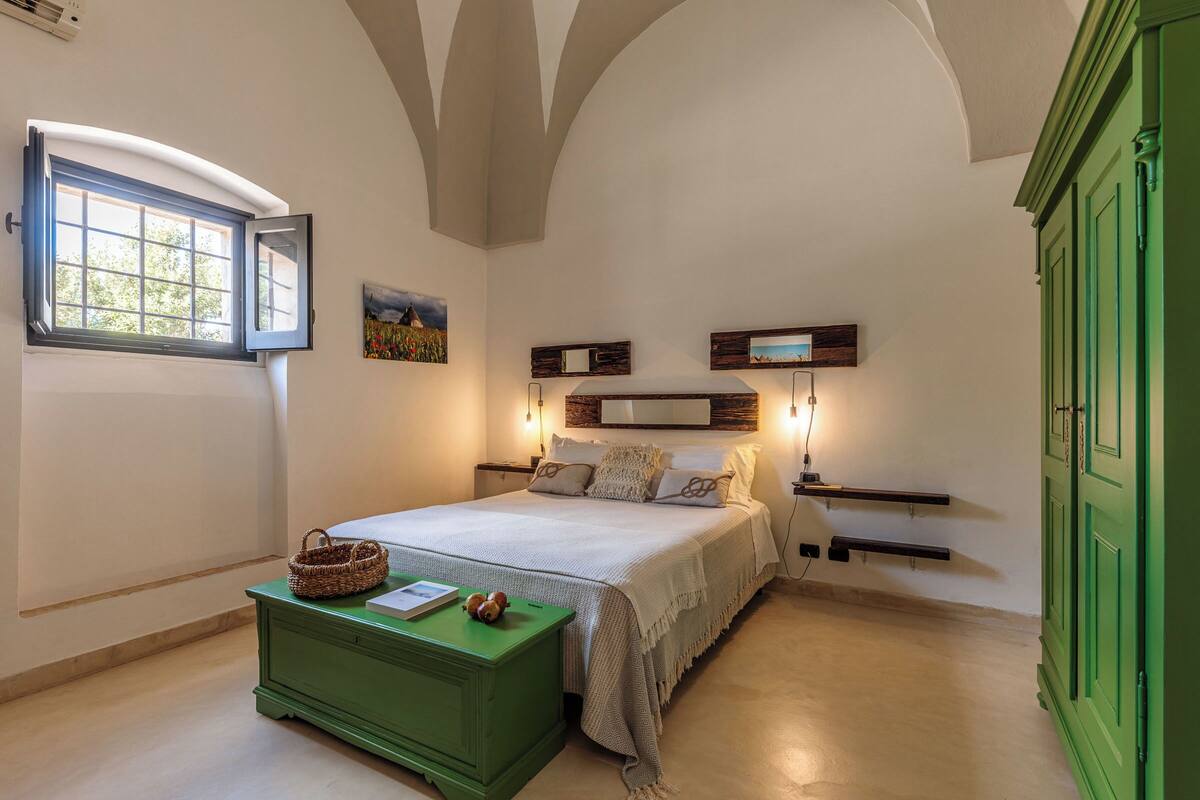 A bedroom with a bed, bedside tables, a green dresser, a window, and wall-mounted shelves.
