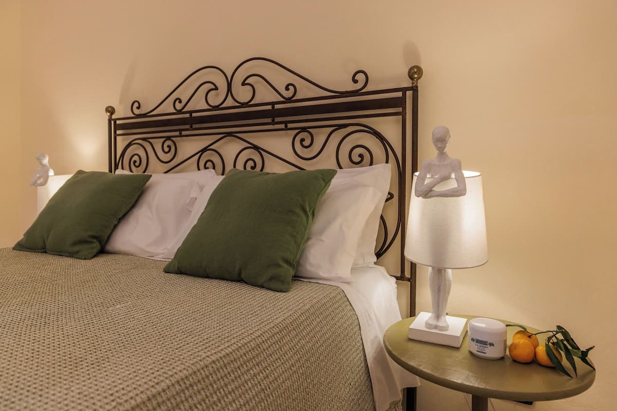 A neatly made bed with green and white pillows, a bedside table with a lamp and a small fruit bowl.