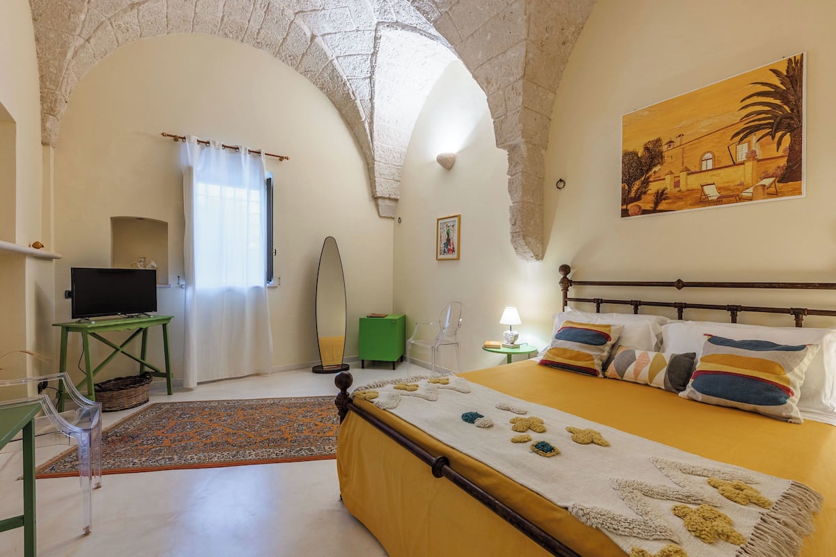 A room with a bed, a television, a green table, a chair, a lamp, and a painting on the wall.