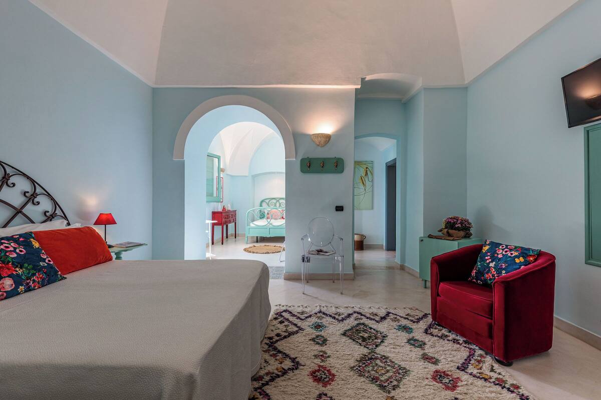 A bedroom with a bed, a red chair, a clear chair, and a small table with a lamp.