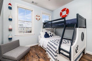 Ein Stockbett mit maritimer Dekoration, ein graues Sofa und ein Blick auf Gebäude durch die Fenster.