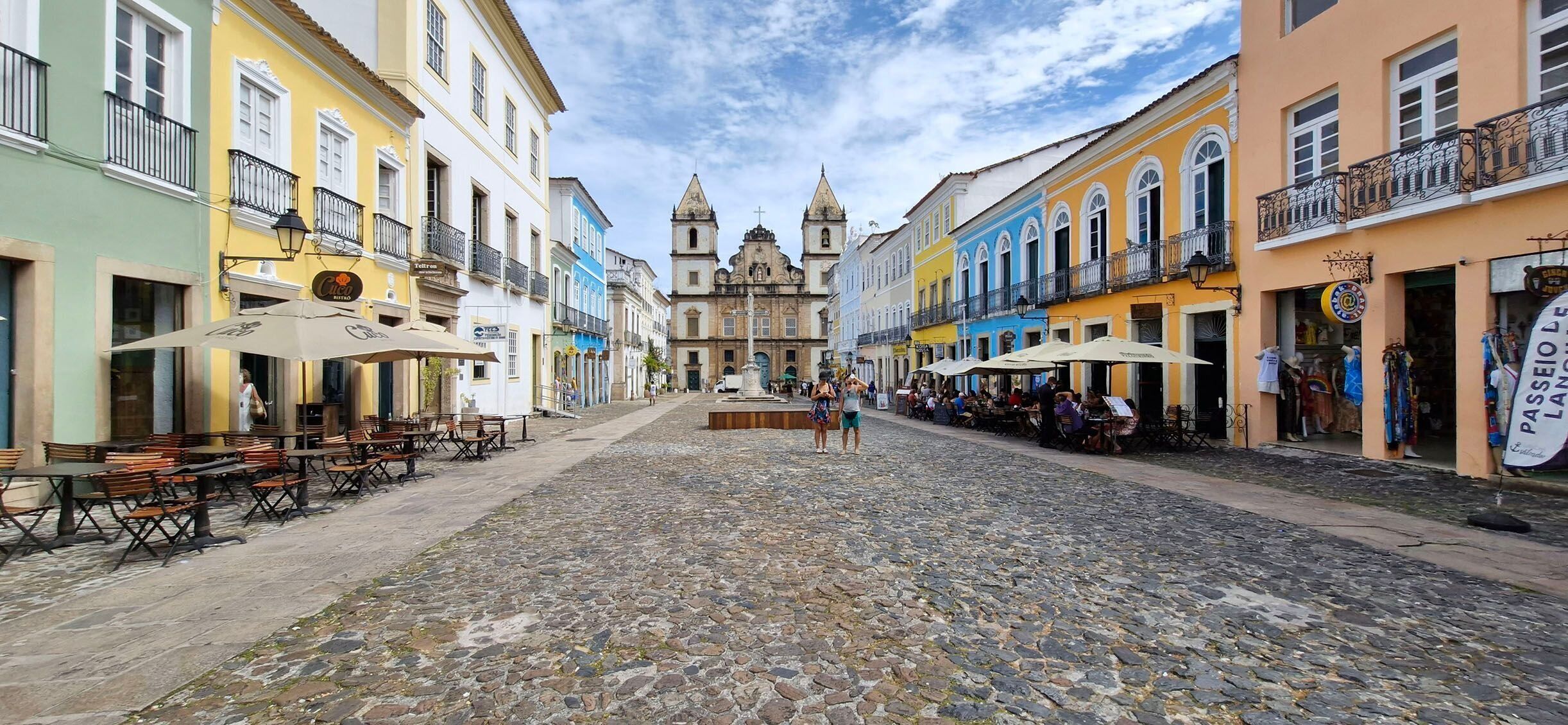A cobblestone street with colorful buildings, outdoor seating, and a central plaza.