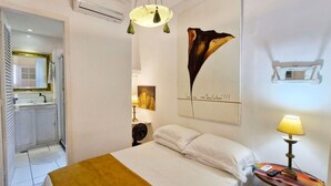 Une chambre d’hôtel avec un lit, une table de chevet avec une lampe, un climatiseur fixé au mur, un miroir et un tableau.