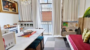 Un espace de vie compact comprenant un coin repas, une petite kitchenette, un lit avec des coussins verts et une grande fenêtre donnant sur un immeuble.