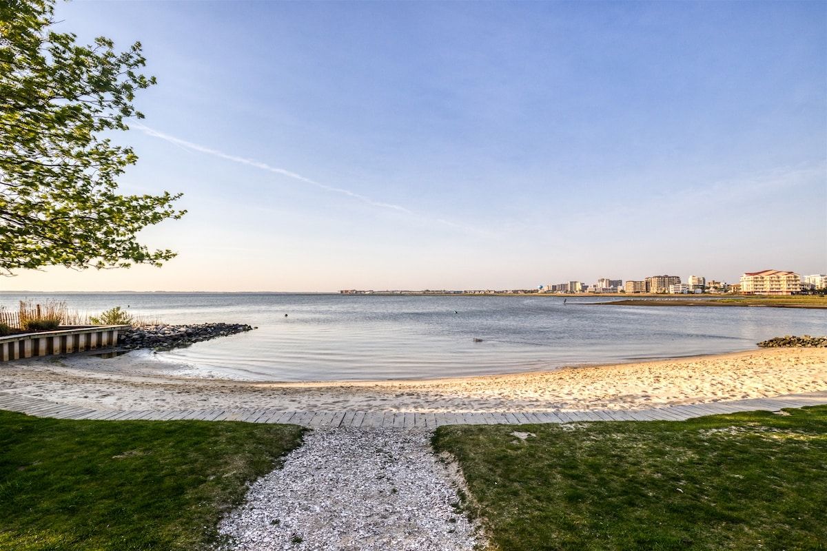 A coastal walkway leading to a beach with a clear view of the ocean and distant buildings.