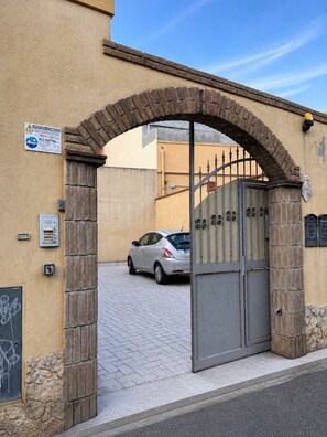 A gated entrance with a brick archway, a car parked inside, and a sign indicating an automatic door. - Su Firindeu Apartment 2 rooms - 15 min from Cagliari and beach - fast Wi-Fi (Monserrato)