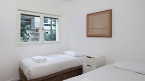 Ein Schlafzimmer mit zwei Betten, einem Fenster mit Blick auf Gebäude und Bäume sowie einem kleinen, weißen Schrank.