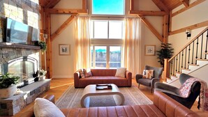 A spacious living room with a stone fireplace, a large window with curtains, a wooden staircase, and a cozy seating area. - Huge Luxury Home, Hot Tub, Movie Theater, Mountain Views (Campton)
