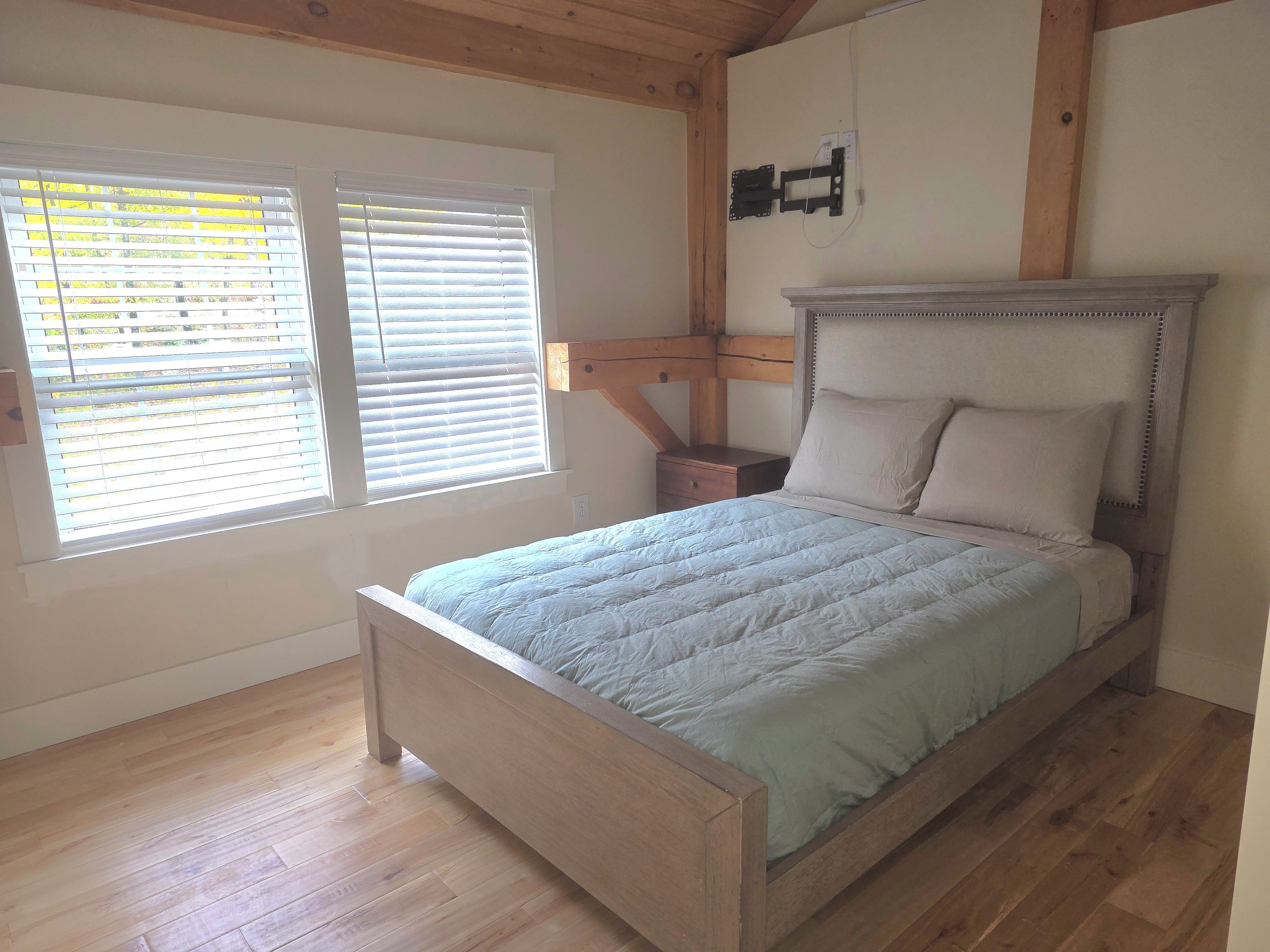 Un dormitorio con cama de madera, una mesita de noche, televisor montado en la pared y una ventana con persianas.
