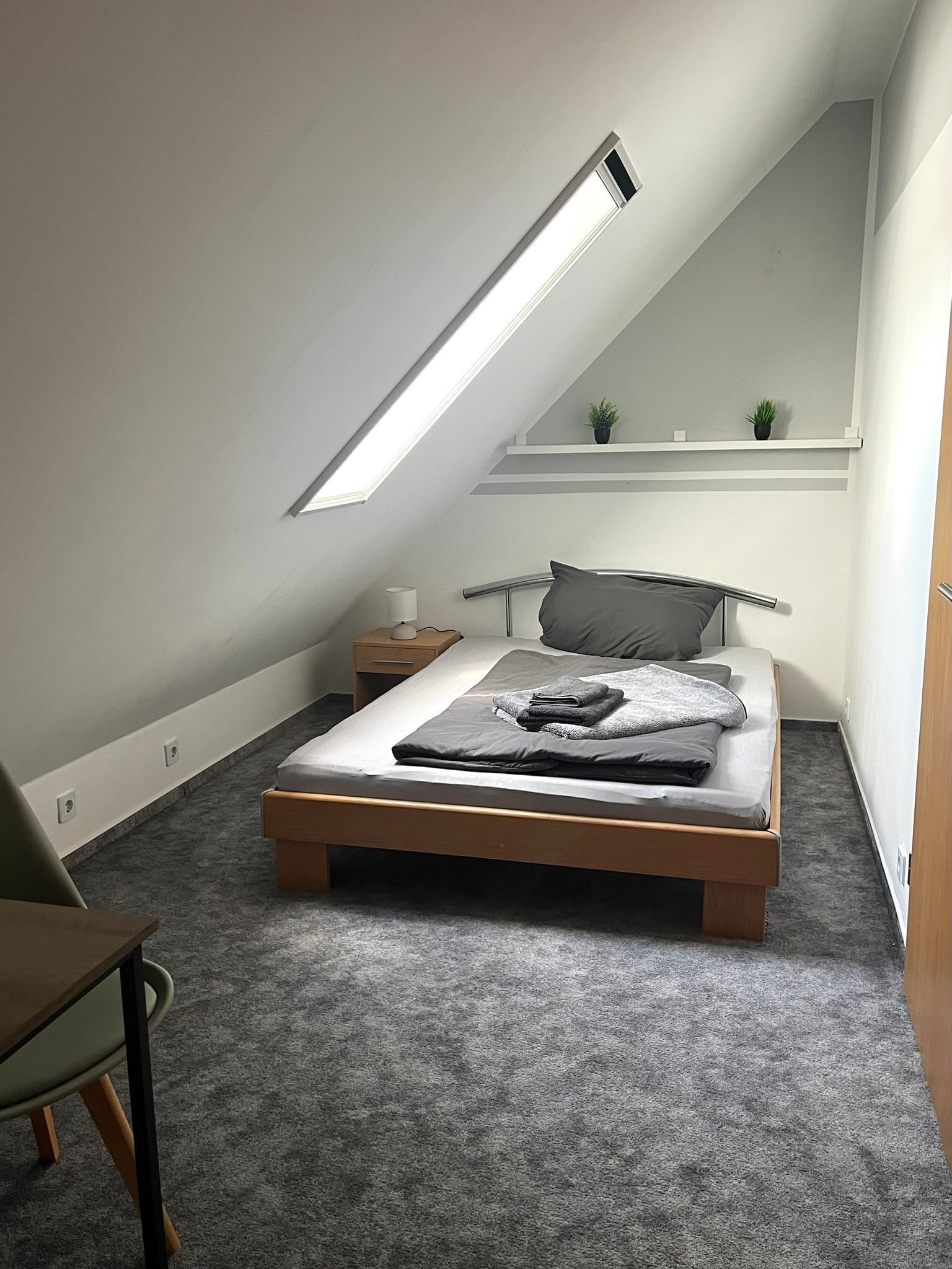 Ein Schlafzimmer mit schrägem Dach, einem Bett, einem Nachttisch, einer Lampe, einem Sessel und einem kleinen Regal mit Pflanzen.