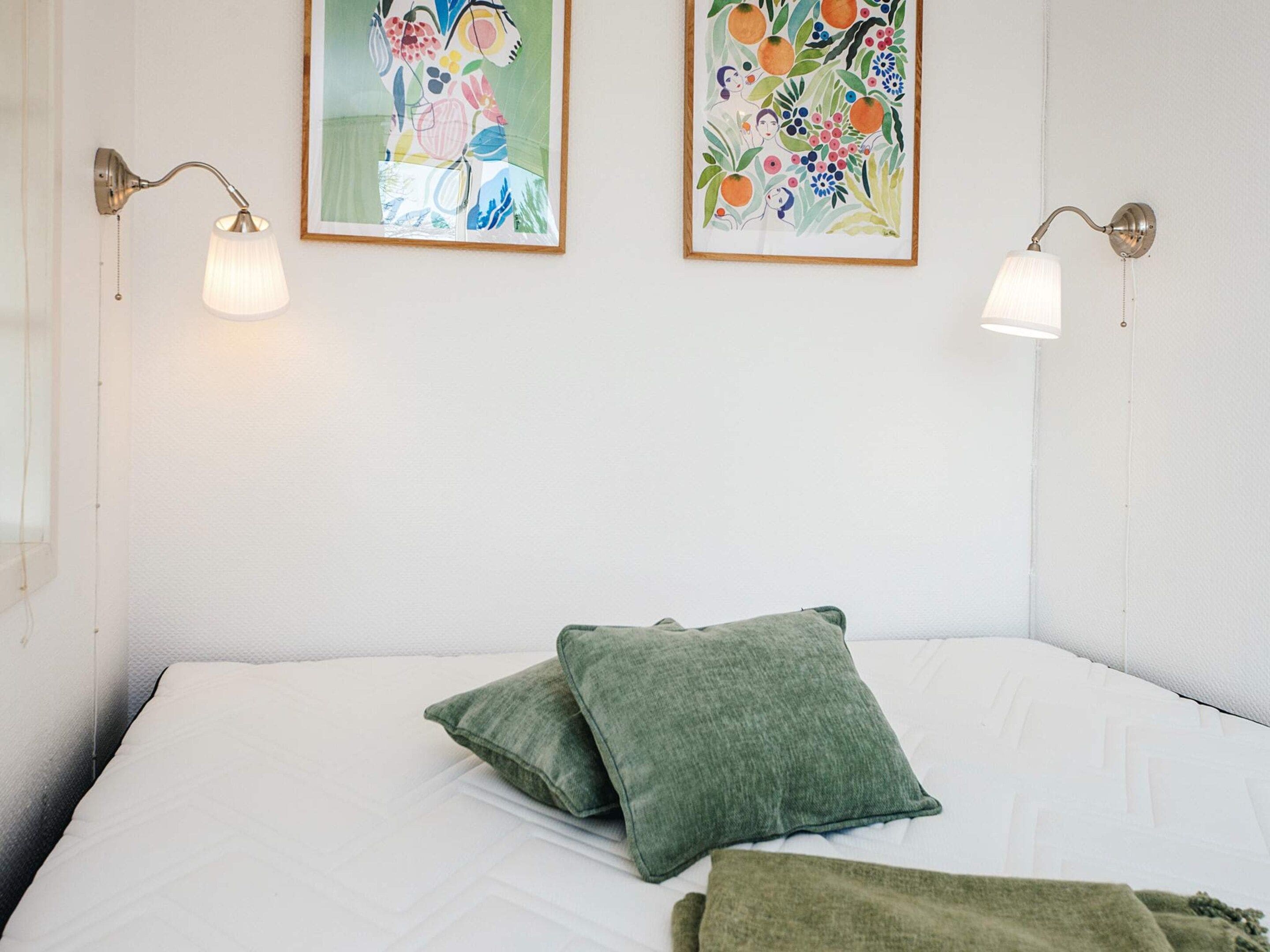 A bedroom with a bed, two wall-mounted lamps, and two framed artworks.