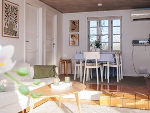 Una habitació amb zona de menjador, un sofà blanc, una taula petita amb espelmes i una finestra que dona a uns arbres.