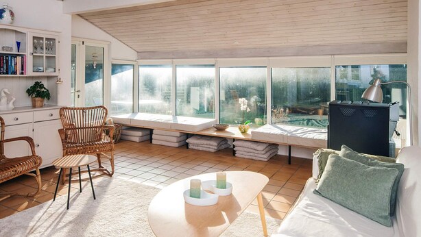 A bright, sunlit room with a large window, a sofa, a table, and a chair. - 6 Person Holiday Home in Juelsminde (Juelsminde)
