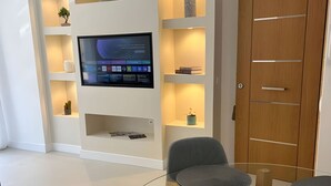 Een moderne woonkamer met een flatscreen televisie aan de muur, een houten garderobe en een glazen salontafel met twee grijze stoelen.