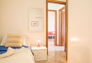 Ein Schlafzimmer mit Bett, Nachttisch und Beistelltisch mit Lampe. Eine Tür führt in einen anderen Raum.