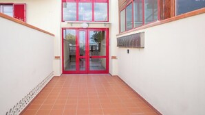 Un corridoio con una porta rossa che dà su una porta in vetro, pavimento piastrellato e una piccola finestra a destra.