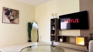 A modern living room with a flat-screen TV displaying 'NETFLIX', a glass coffee table, a painting on the wall, and a plant. - Spacious 2BR Apartment ,3 Bathrooms ,Near Mall (Abu Dhabi)