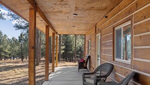 A wooden cabin with a covered porch, wicker chairs, and a view of the surrounding forest.
