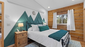A bedroom with a bed, a nightstand, a wooden headboard, a window with curtains, and a wall with a mountain design.