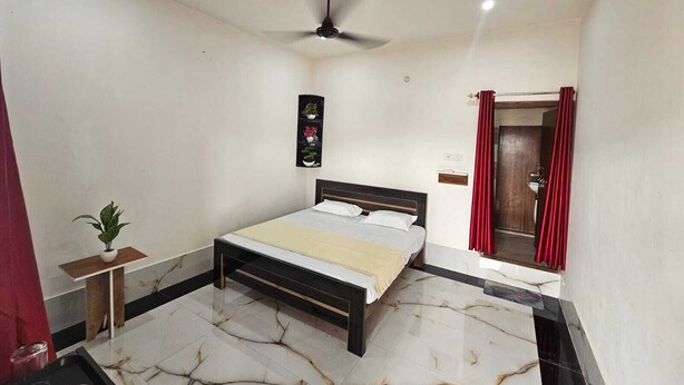 A bedroom with a bed, a ceiling fan, a small table with a plant, and a red curtain. - Relax With Ramayana (Ayodhya)