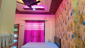 A bedroom with a bed, a ceiling fan, a window with pink curtains, and a wall with a peacock pattern. - Relax With Ramayana (Ayodhya)