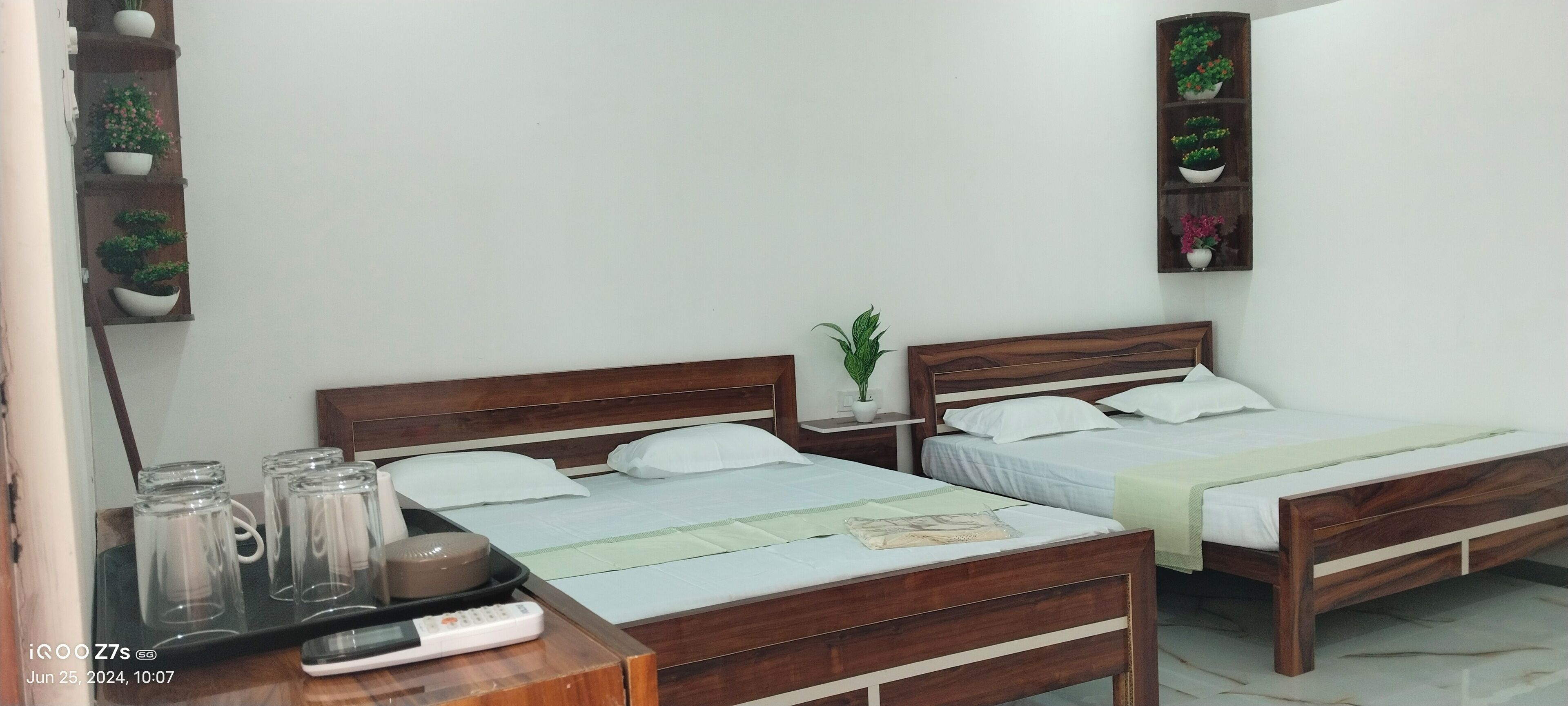 A hotel room with two wooden beds, a bedside table with a lamp, a small potted plant, and a shelf with decorative items.
