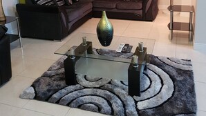Een woonkamer met een hoekbank, een glazen salontafel, een vloerlamp en een kamerkleed met een patroon.