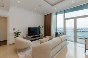 A modern living room with a flat-screen TV, beige sofas, a coffee table, and a large window offering a city view. - Prime 3BR w Beach Access at the Palm (Dubai)